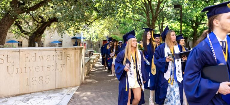 A crowd of happy students in graduation gowns gather outside in front of the fountain at St. Edward's University