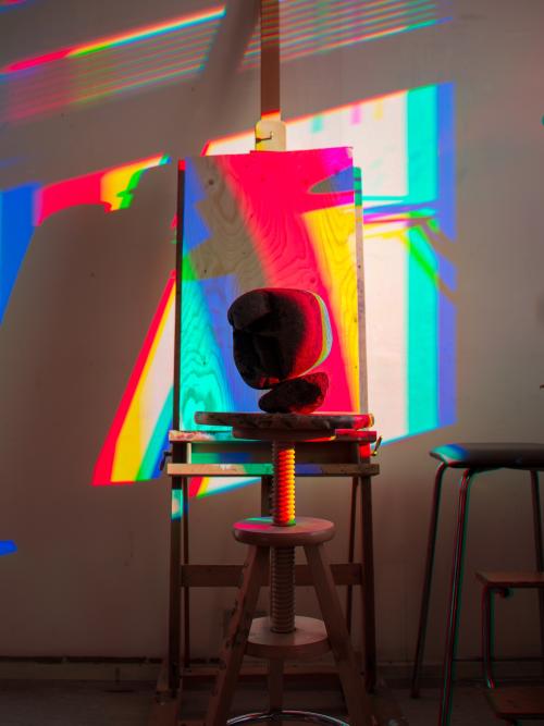 A colorful photograph of a large rock in front of an easel, with primary colors. Photo by artist Glenn Twiggs, entitled "Color Time Still Life #2, Old School Residency", 2024