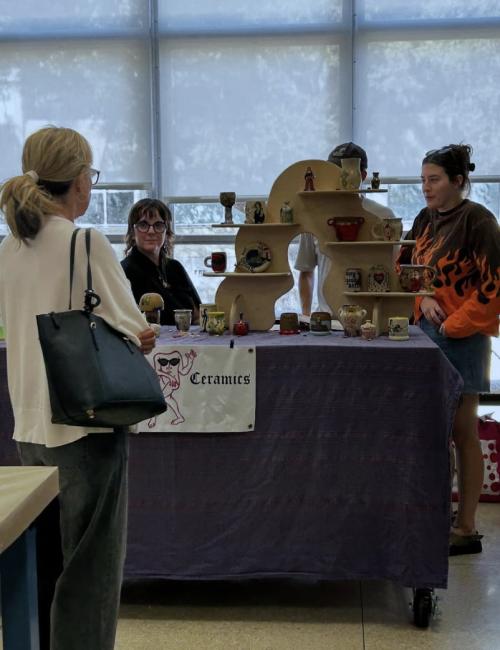 2 people standing behind a table at an art fair, one person standing in front of the table, talking to them.