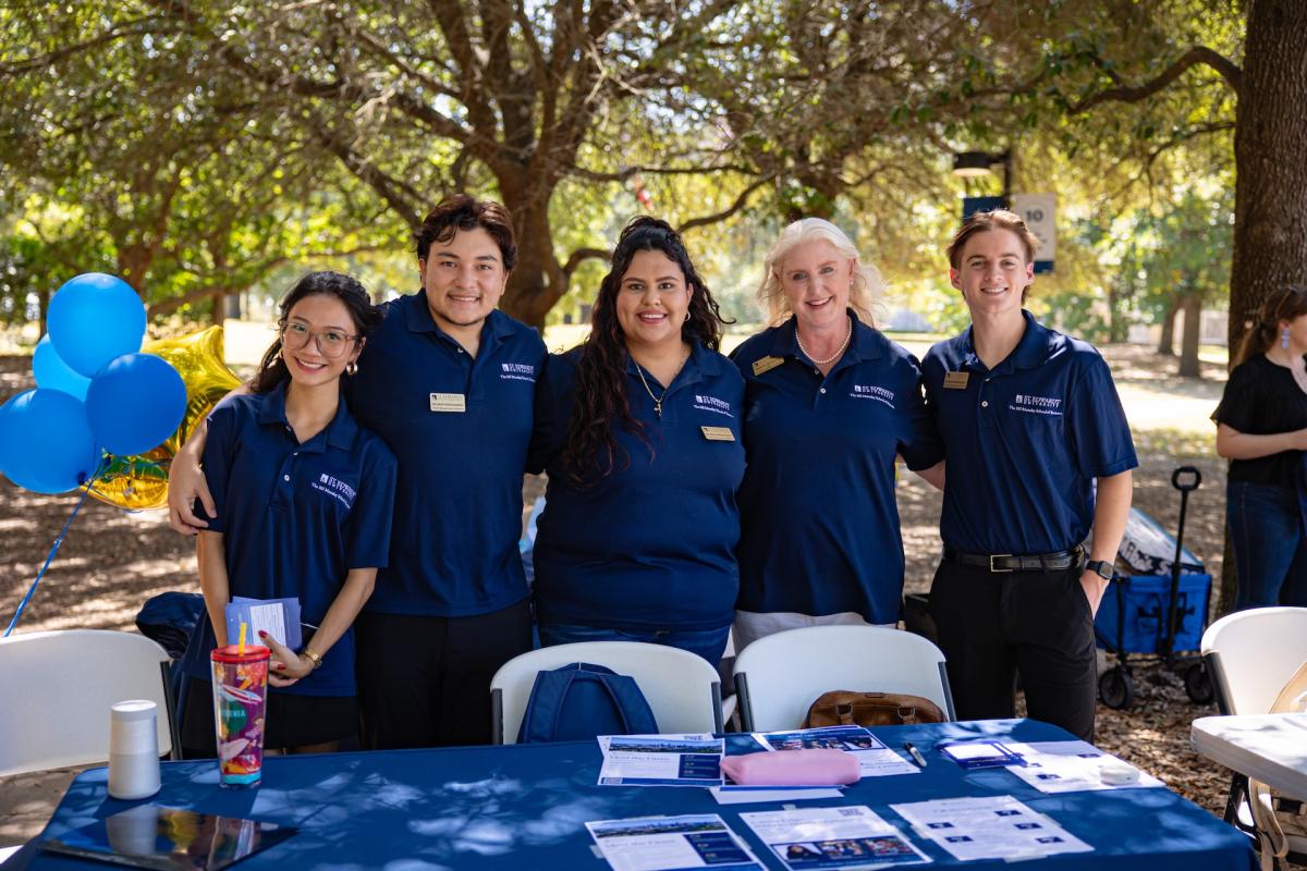 Five people wearing navy blue photos stand behind a table outside and smile for a photo.