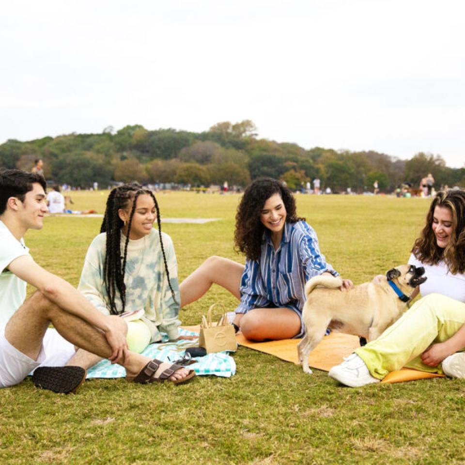 Taylor Huey, second from left, sits with three friends on colorful towels and yoga mats in Zilker Park and they pet a dog.
