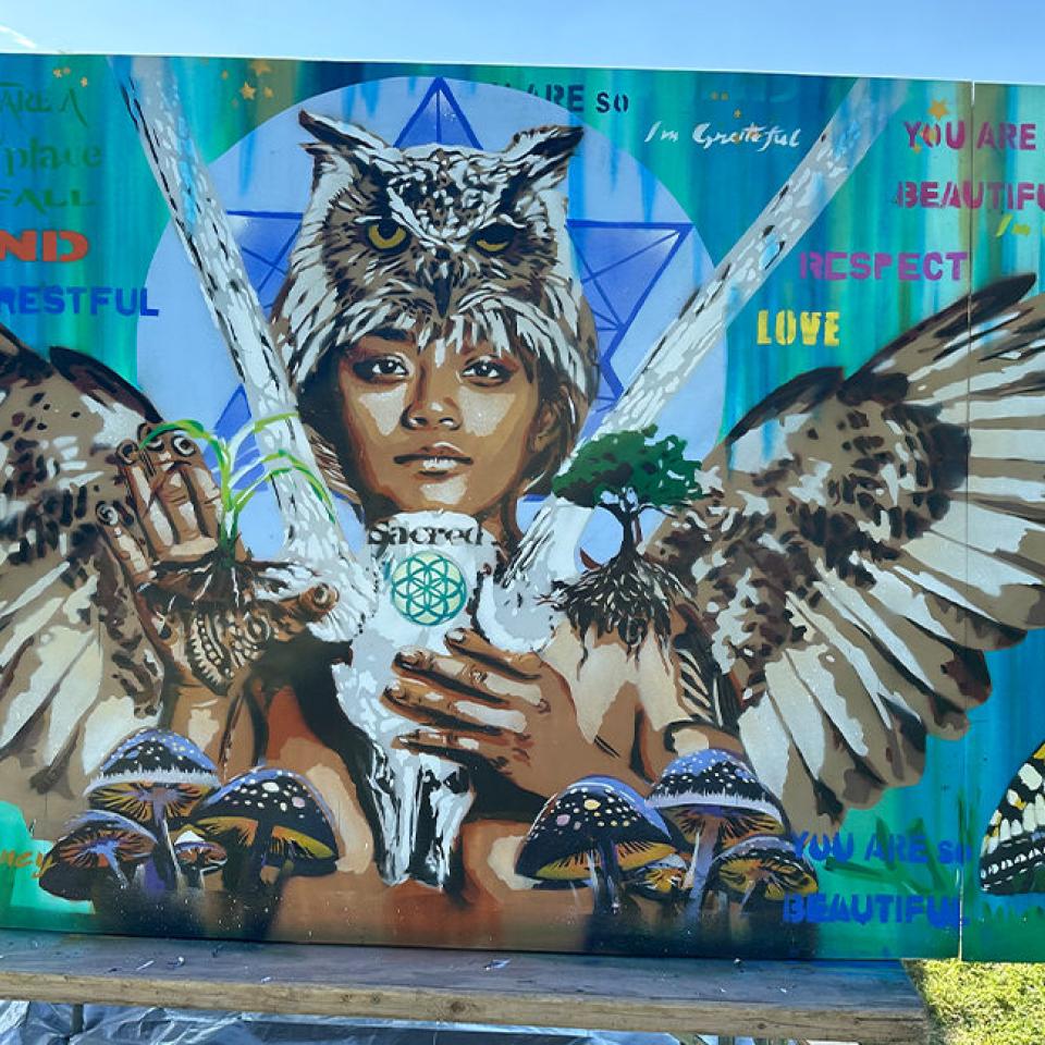 The image is a vibrant mural featuring a young person's face with an owl perched on their head. Their hands hold various elements: a plant, a tree, and a white object with the word "sacred" and a geometric design. The mural incorporates words like "kind," "restful," "love," "respectful," and "beautiful," along with phrases like "you are" and "I'm grateful." The background features a blue and green gradient with geometric shapes, creating a theme of nature, wisdom, and positive affirmations.