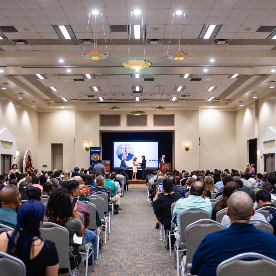 A group of people seated watching a presentation given on stage in the Mabee Ballrooms