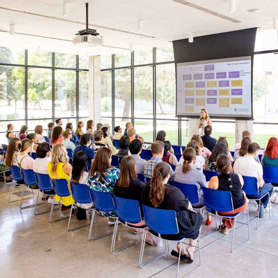 A group of people is seated in chairs in St. Andre Multipurpose Room listening to a speech. 