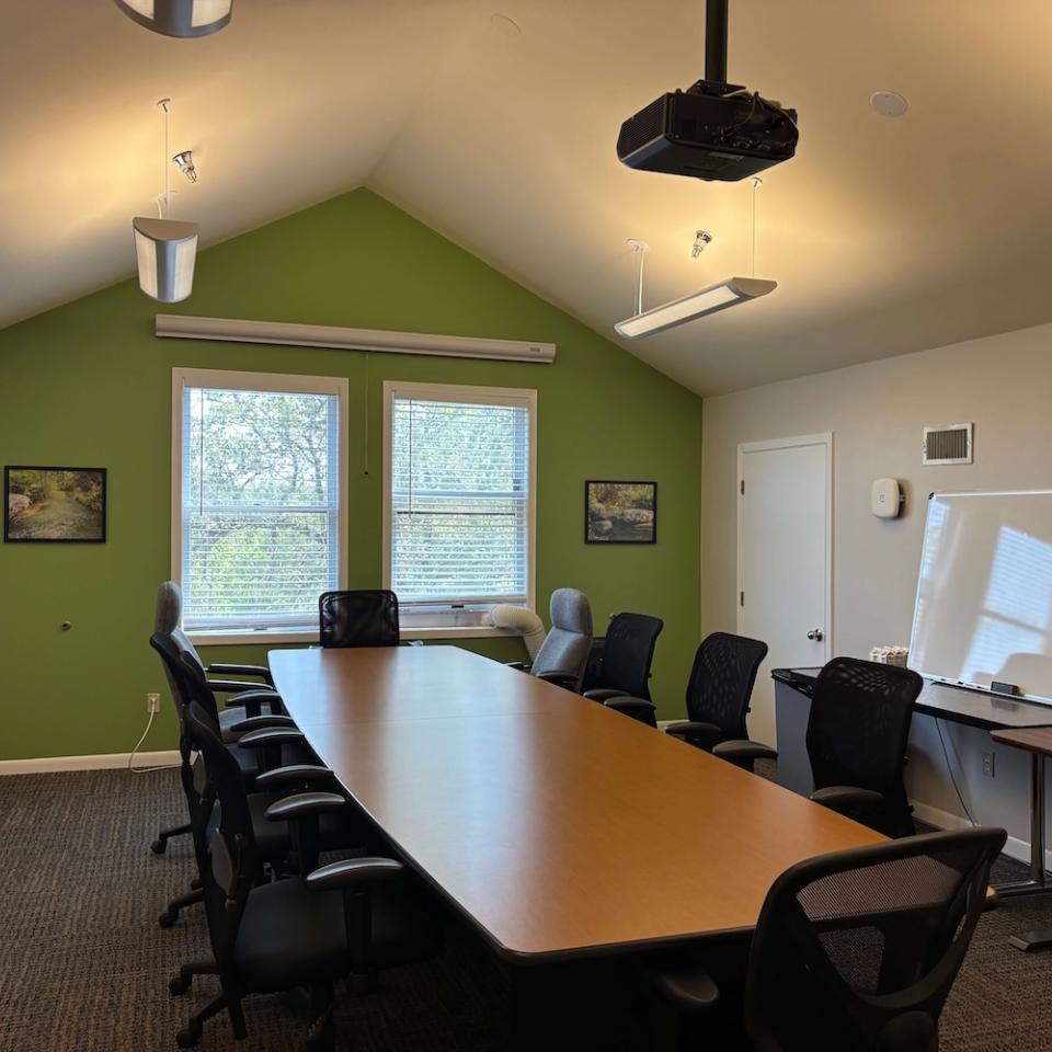  A conference room with a long wooden table, nine black chairs, a bright green accent wall with two windows, and a projector mounted on the vaulted ceiling. The room also has two framed pictures and a whiteboard.