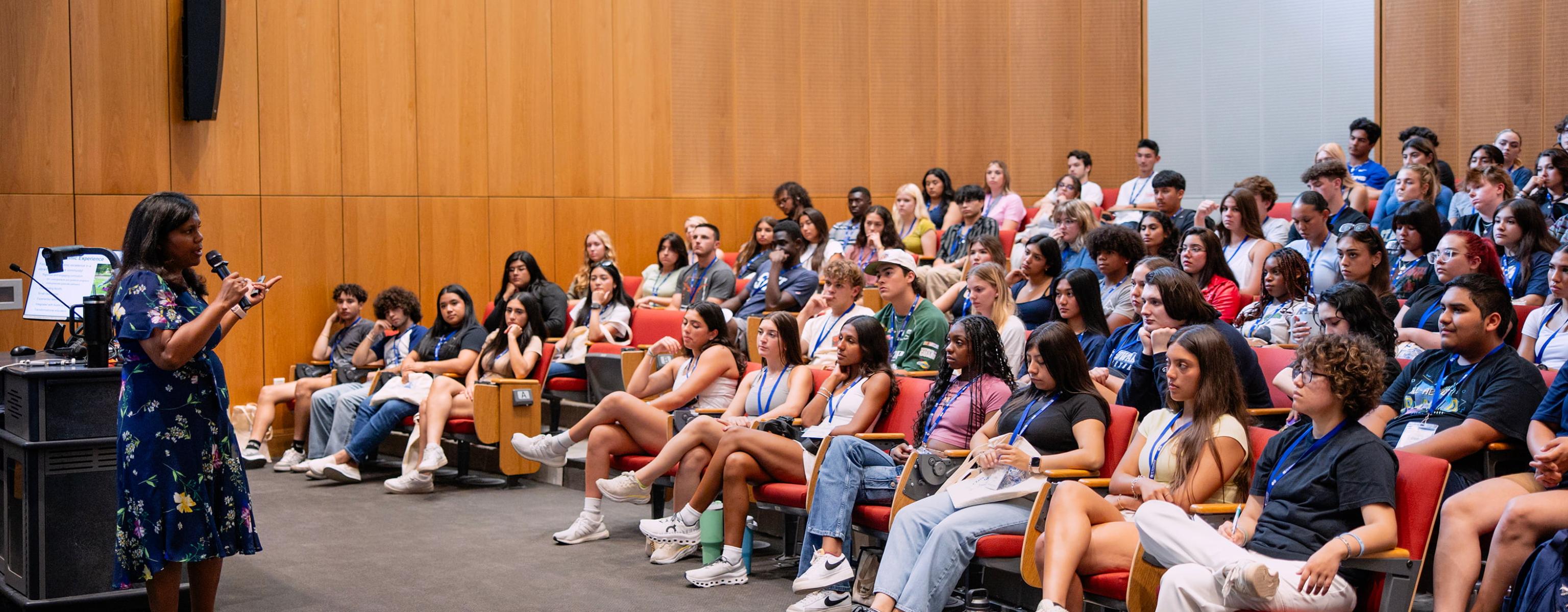 A speaker addressing a group of attentive students seated in an auditorium. The atmosphere reflects an educational event, with students wearing name badges and focused on the presentation.