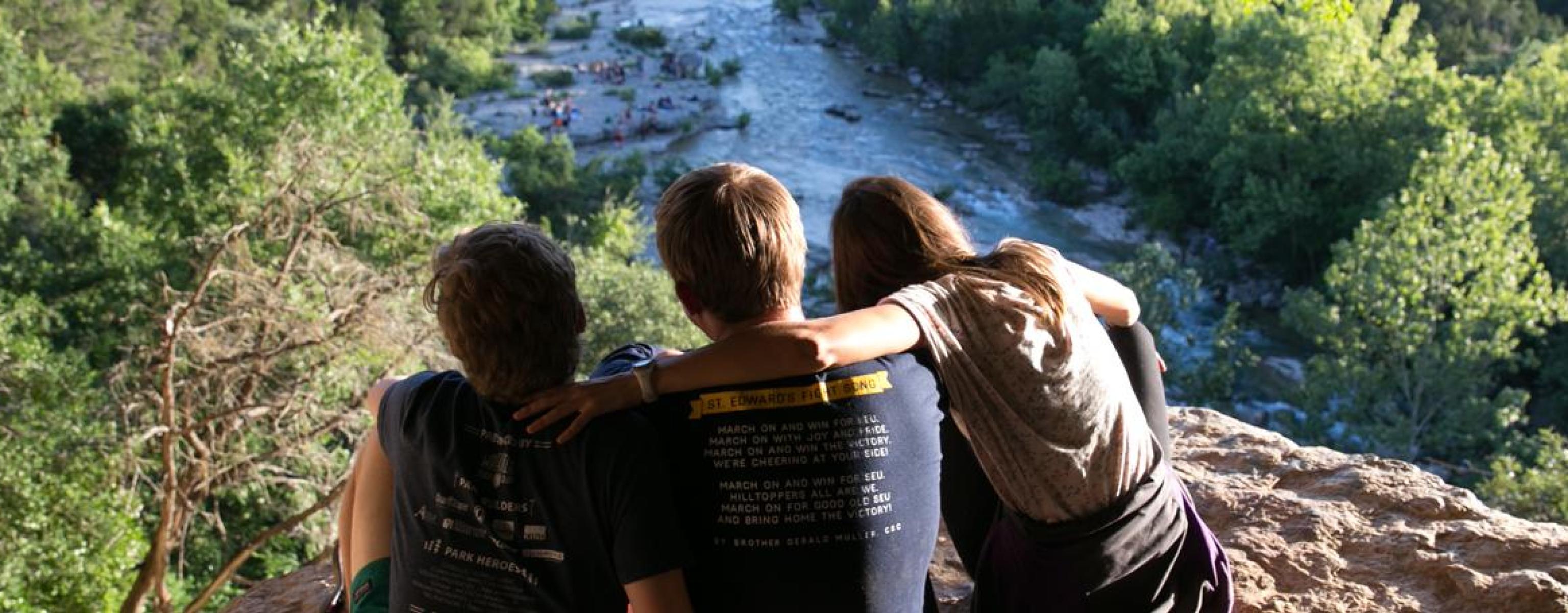 The image shows three friends sitting on a rocky overlook, gazing at a lush, green landscape with a river flowing below. One person has their arm around another in a relaxed and friendly gesture. The background is filled with trees and people enjoying the water, creating a serene and adventurous outdoor setting. Sunlight filters through the leaves, casting a warm glow over the scene.