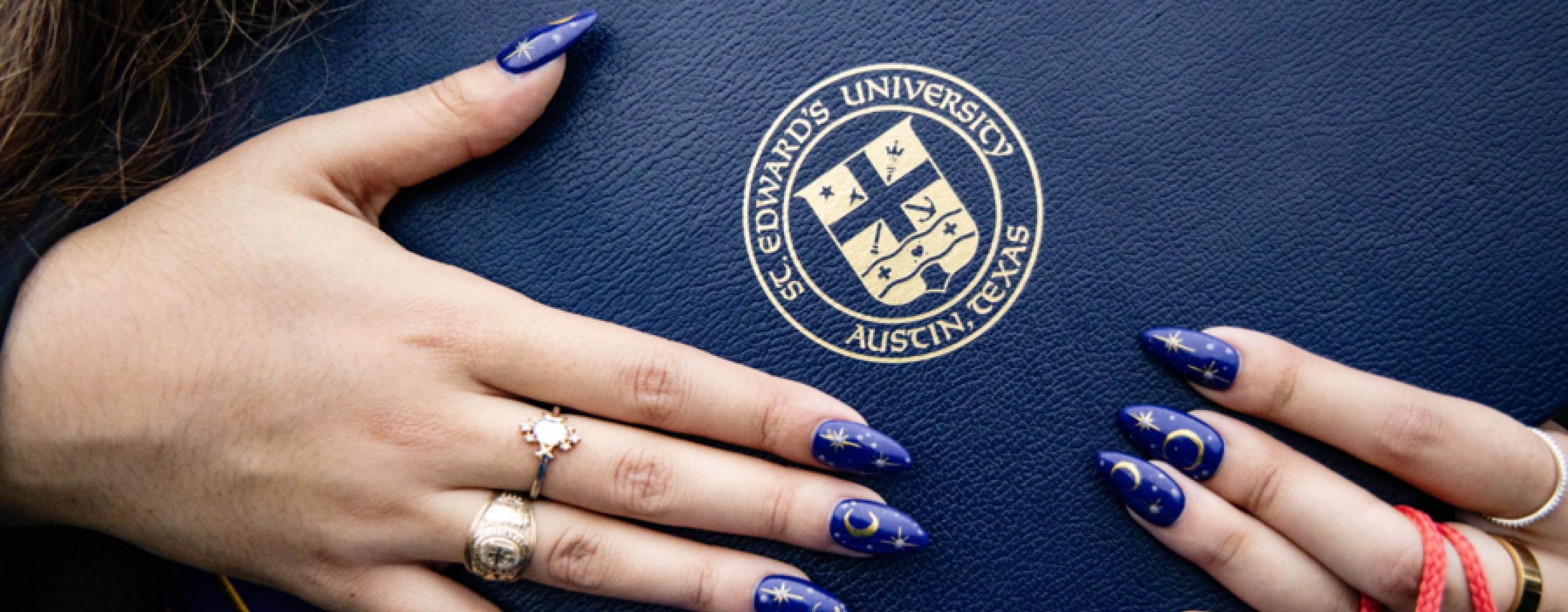 a 2024 graduate holds their diploma cover, showing off the St. Edward's seal and beautiful nails painted with the moon and stars