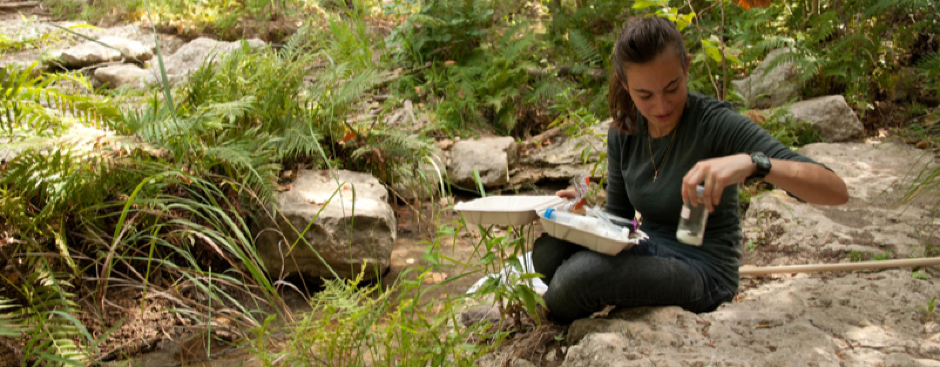 A woman with a notebook sitting in the woods by a creek