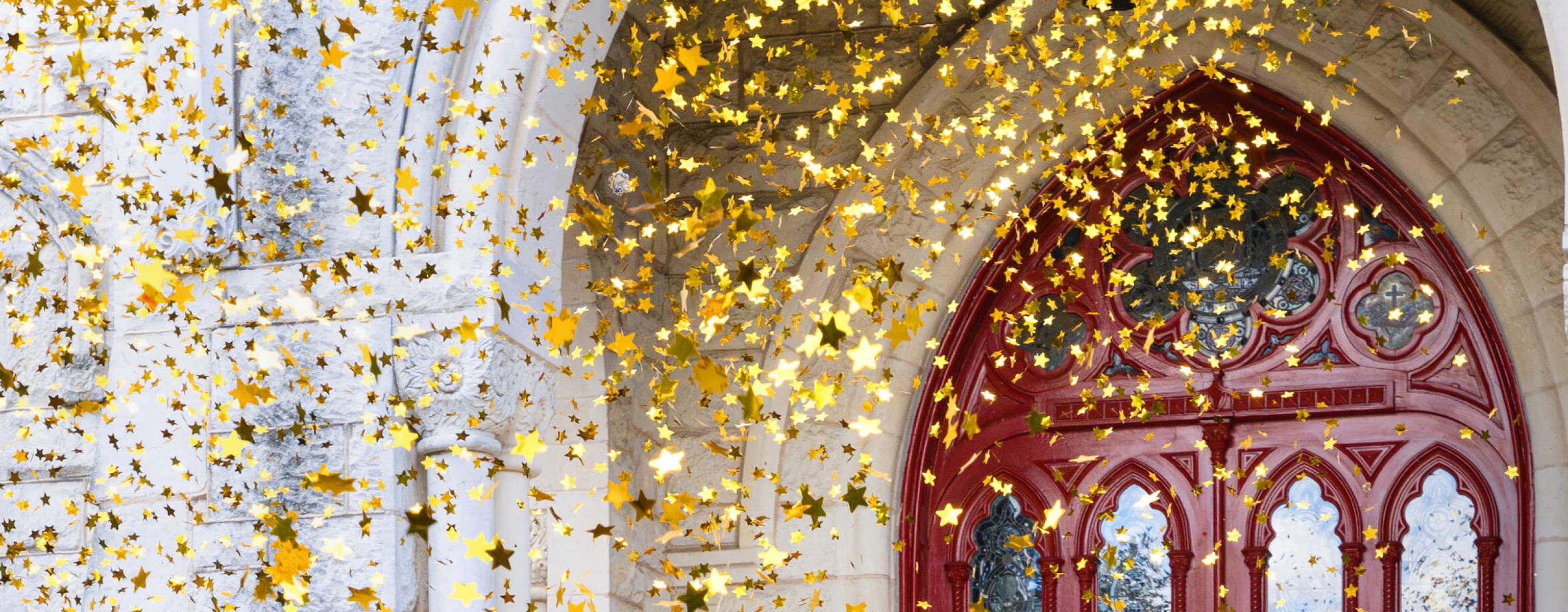 Gold star confetti is shown in the air in front of the red doors of Main Building