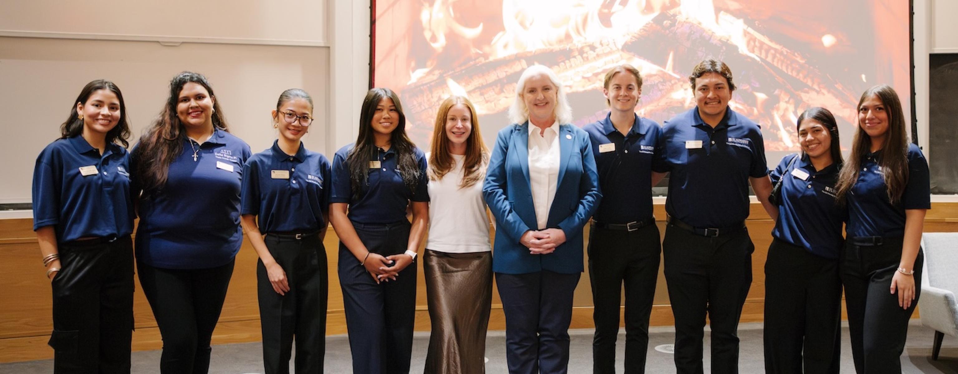 A group of college students smile for a photo with two women in front of a fireplace background. 