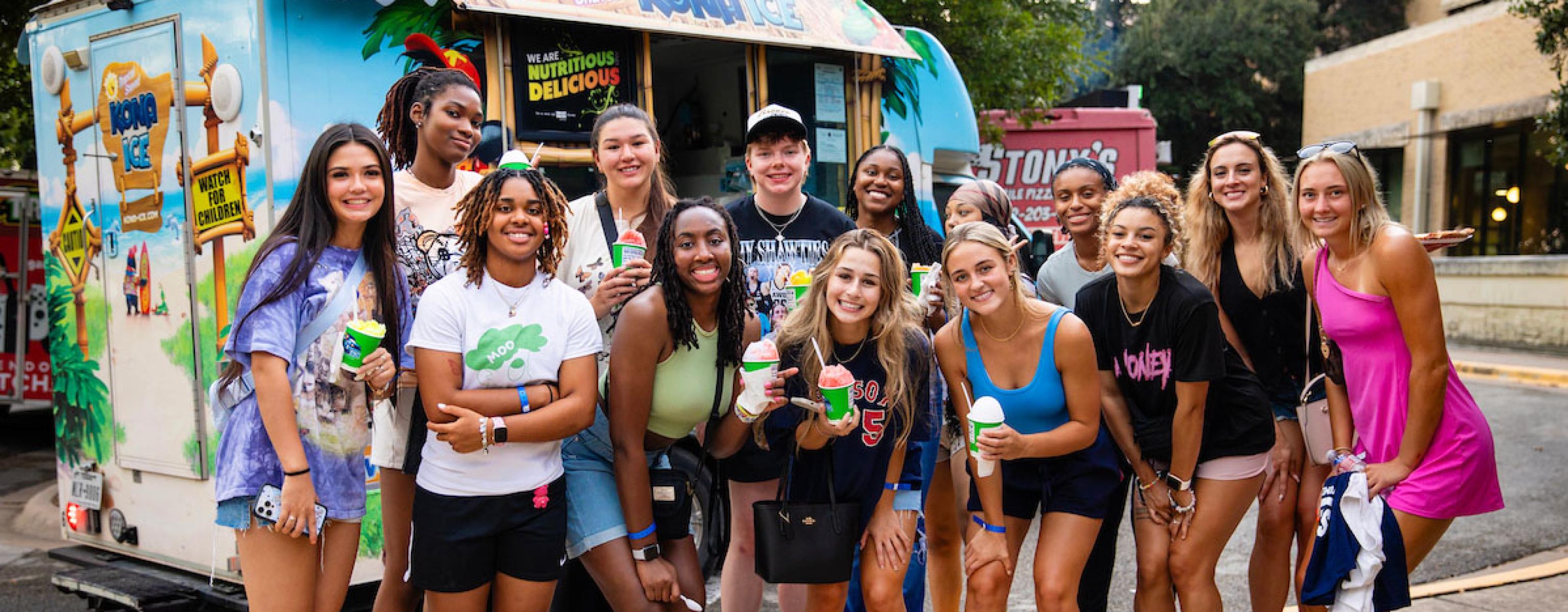 A large group of smiling students posing together on a sidewalk in front of a colorful Kona Ice shaved ice truck at an outdoor event. Several people in the group are holding up bright green cups filled with shaved ice.