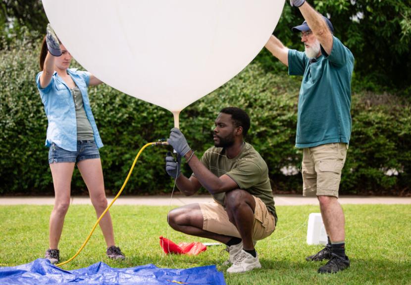 Three people are outside on a grassy area on the St. Edward's University campus, working together to inflate a large white weather balloon using a yellow hose attached to the neck.