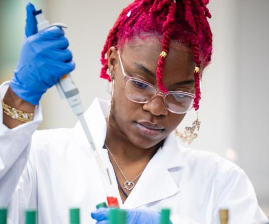 A focused student with bright pink twisted hair and a white lab coat uses a micropipette to work with samples in a microbiology lab. She wears blue gloves and safety glasses while working over a bench filled with test tubes and petri dishes.