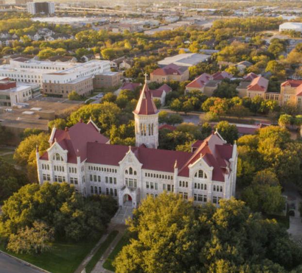 The image shows an aerial view of a university campus with a prominent, large building featuring red roofs and a central tower surrounded by lush green trees. Other buildings with similar red roofs are visible throughout the campus. The surrounding area includes a mix of greenery and urban development. The sky is clear, and the overall atmosphere appears calm and expansive, indicating a spacious and well-planned campus layout.