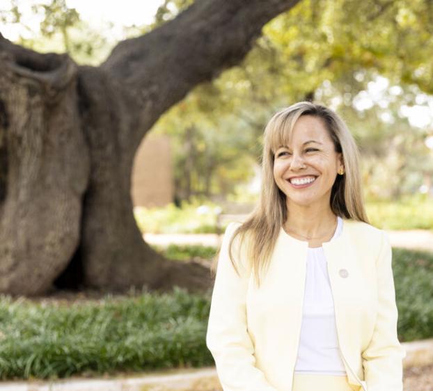 The image features an individual standing in front of a large tree with a thick trunk and sprawling branches. The individual is wearing a light-colored blazer, and the setting appears to be a park or garden with greenery in the background.