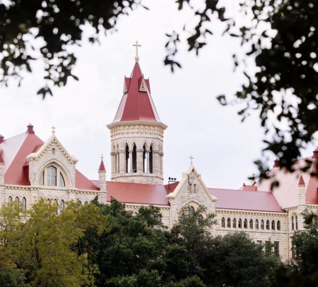 The image features a grand, historic building with a red roof and a prominent central tower topped with a cross. The architecture is reminiscent of a university building, with pointed arches, large windows, and intricate stonework. The building is partially framed by tree branches, adding a natural element to the composition. The overall scene suggests a peaceful, stately environment, likely on a college campus.