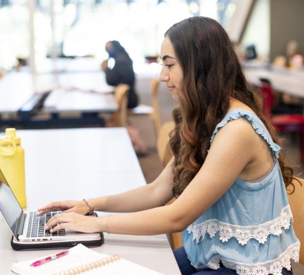 The image shows a young woman with long, wavy brown hair working on a laptop in a spacious, well-lit study area. She wears a light blue sleeveless top with white lace trim. A yellow water bottle, a pink pen, and a notebook are on the table. Other students are visible studying in the background.