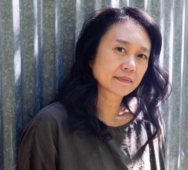 A woman with wavy dark hair stands against a corrugated metal wall, gazing calmly at the camera. She wears a loose, dark-colored top, and soft sunlight highlights her face and hair.