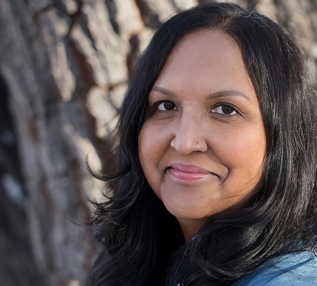 A woman with long, dark hair stands outdoors in front of a tree trunk, smiling softly at the camera. She wears a nose ring and a denim jacket, with natural light highlighting her face and hair.
