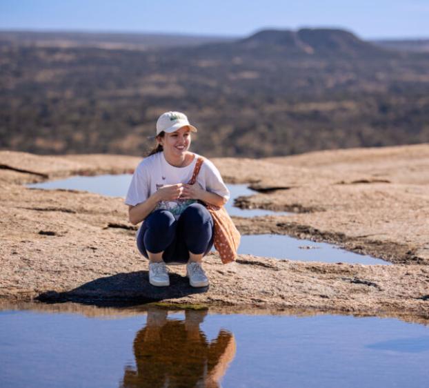 A St. Edward's University student sits crouched in front of water at Enchanted Rock Natural State Natural Area outside of Austin with a rocky horizon and clear blue sky in the background