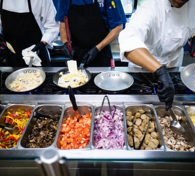 Chefs in black gloves and aprons prepare food at a cooking station with skillets on burners. In front of them are trays filled with ingredients including bell peppers, mushrooms, tomatoes, onions, meatballs, chicken, and pasta, ready to be added to the dishes they are making.