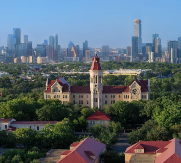 An aerial view of Main Building, with the downtown Austin skyline in the background. Other campus buildings' roofs can be seen, as well as lush green trees.