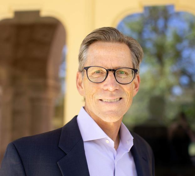 A professional headshot of a smiling man with short, styled gray hair and glasses. He is wearing a dark navy blazer over a light purple button-down shirt. The background shows a blurred outdoor architectural setting with arches and greenery