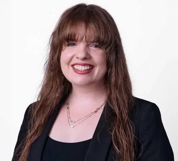 A professional headshot of a smiling woman with long brown hair and bangs. She is wearing a black blazer over a black top, accented with a gold necklace. The background is a clean, solid white.