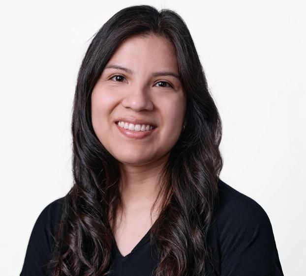 A professional studio headshot of a woman with long, dark wavy hair and a friendly smile. She has a fair-to-medium complexion and is wearing a black V-neck top. The background is a clean, solid white with soft lighting that highlights her features.