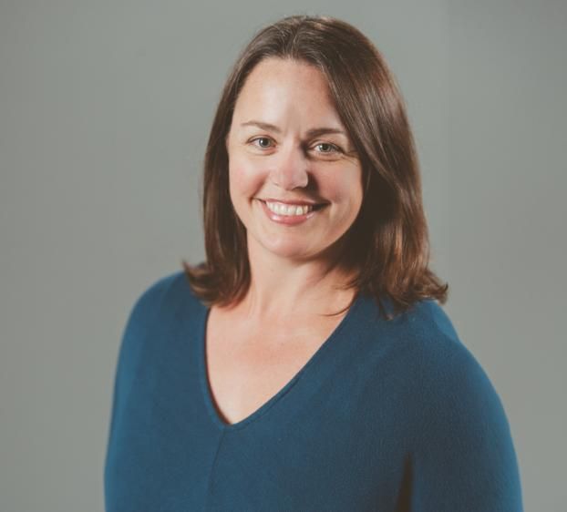 A professional headshot of a smiling woman with medium-length brown hair and light-colored eyes. She is wearing a dark teal V-neck sweater and is positioned against a solid, neutral gray background. The lighting is soft and even, highlighting her friendly expression.