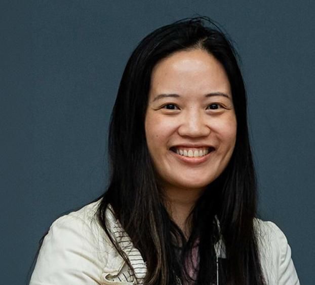 Headshot of a smiling woman with long, dark hair. She is wearing a light-colored jacket over a dark top. She has a warm expression and is looking directly at the camera. The background is a solid, neutral gray-blue.