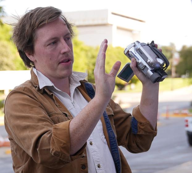 A man with light brown hair and freckles stands outdoors, holding a silver handheld camcorder. He is wearing a brown jacket over a white button-down shirt with a blue strap across his shoulder. He has one hand raised near the camera as if adjusting a shot or explaining something. The background is softly blurred, showing a street, greenery, and a building under bright daylight.