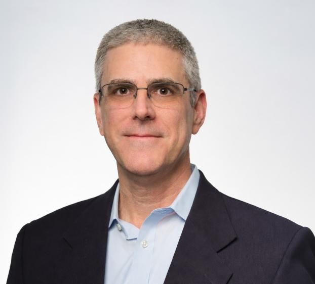 A professional headshot of a man with short, salt-and-pepper hair and glasses. He is smiling subtly and wearing a light blue button-down shirt under a dark navy blazer. The background is a solid, neutral light gray. The lighting is bright and even, focusing on his face from the chest up.