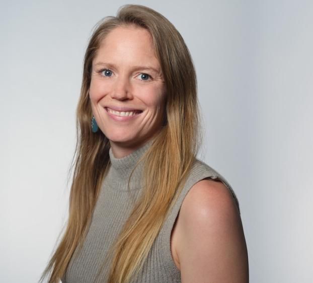 A professional headshot of a woman with long blonde hair, smiling at the camera. She is wearing a grey sleeveless turtleneck sweater and a turquoise earring. The background is a clean, neutral light grey. The lighting is bright and even, highlighting her facial features.