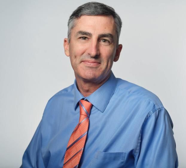 A head-and-shoulders portrait of a man with short, salt-and-pepper hair. He is smiling slightly and looking toward the camera against a plain, light-gray background. He wears a blue, long-sleeved button-down dress shirt and an orange necktie with thin blue diagonal stripes. The lighting is bright and even, highlighting his professional attire and friendly expression.