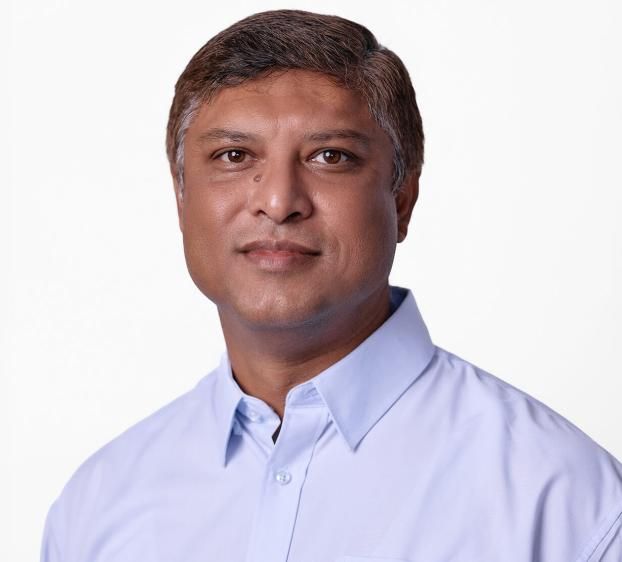 A headshot of a man with short, salt-and-pepper hair and brown eyes, looking directly at the camera with a neutral, professional expression. He has a light brown complexion and a small mole under his right eye. He is wearing a light blue, button-down collared shirt. The background is solid white, and the lighting is bright and even.