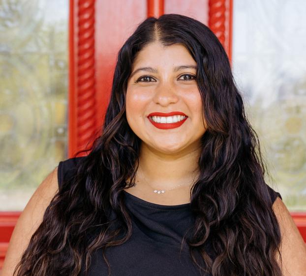 A smiling woman with long, wavy dark hair and bright red lipstick. She is wearing a black top and a delicate necklace with three small charms. She is positioned in front of a vibrant red door with decorative glass panels and intricate wood carving.