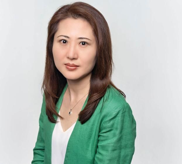 A professional headshot of a woman with shoulder-length brown hair and a slight smile. She is wearing a green linen blazer over a white top and a delicate silver necklace with a dark pendant. The background is a clean, solid light grey. The lighting is bright and even, highlighting her face clearly.