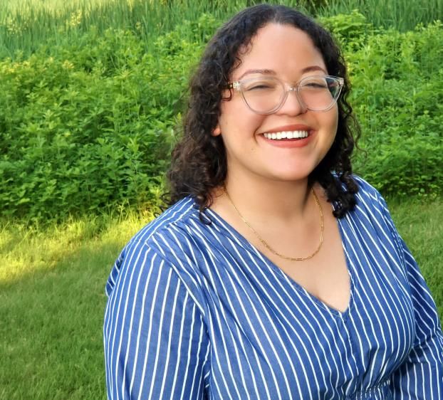 A smiling woman with curly brown hair and clear-framed glasses stands outdoors. She is wearing a blue and white striped V-neck top and a thin gold chain necklace. The background is a lush green field of grass and foliage under bright, natural light.