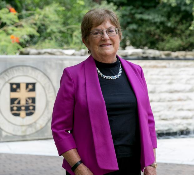 A medium shot of a woman with short brown hair and glasses, smiling in front of a stone wall. She wears a vibrant magenta blazer over a black top and a silver statement necklace. To her left, a large stone seal featuring a crest is visible. The background includes lush green foliage and a stone water feature, creating a professional yet bright outdoor setting.