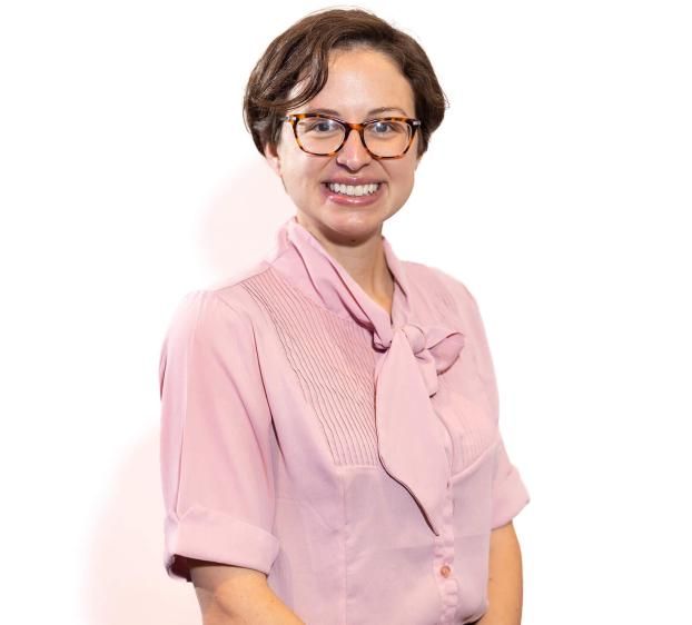 A professional headshot of a smiling woman with short brown hair and tortoiseshell glasses. She is wearing a light pink, short-sleeved blouse with a pussy-bow neckline and pleated detailing on the chest. She is positioned against a plain, brightly lit white background. The shot is framed from the waist up.