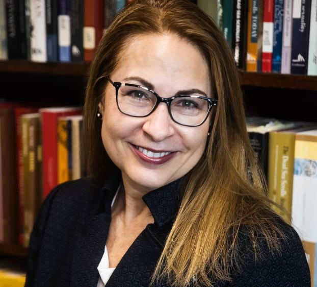 A headshot of a smiling woman with long brown hair and glasses, wearing a dark textured blazer. She is positioned in front of a bookshelf filled with academic books. She has a warm expression and is looking directly at the camera.