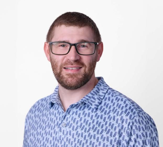 A head-and-shoulders portrait of a man with short brown hair and a groomed beard, wearing black rectangular glasses. He is smiling and wearing a blue short-sleeved button-down shirt featuring a repeating purple floral pattern. The background is a clean, solid white.