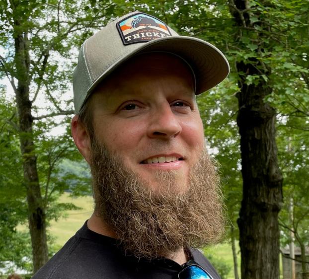 A close-up, slightly angled portrait of a smiling man with a full, light brown beard and mustache. He is wearing a black t-shirt and a grey and tan trucker hat featuring a "Thicket" patch. Sunglasses are tucked into his shirt collar. The background is a soft-focus outdoor setting with lush green trees and a field under bright, natural light.