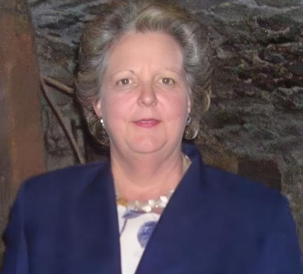 A woman with fair skin and short, styled light-brown hair. She is wearing a dark blue blazer over a floral patterned top and a light-colored necklace. She has a neutral expression and is looking directly at the camera. The background appears to be a textured, dark stone wall or an outdoor setting.