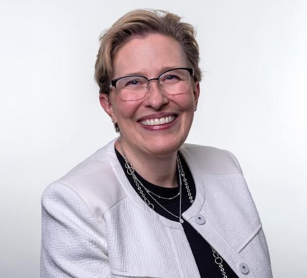 A professional headshot of a smiling woman with short blonde hair and glasses. She wears a white textured blazer over a black top, accented by layered silver necklaces. The background is a clean, solid light gray. Her expression is warm and approachable.