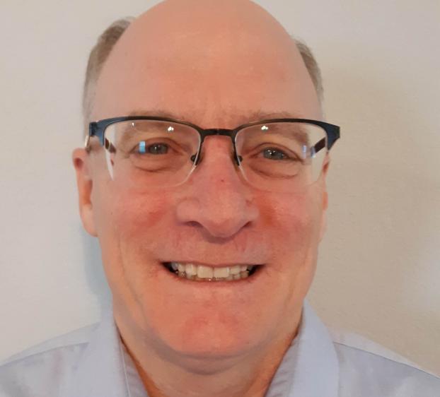 A close-up portrait of a smiling man. He is wearing black-rimmed glasses and a light blue button-down shirt. The background is a plain, off-white wall. The lighting is soft and frontal, creating a clear and friendly professional headshot.
