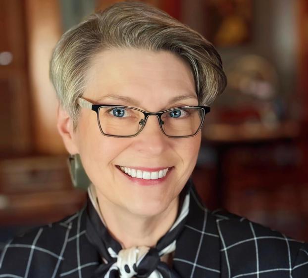 A headshot of a smiling woman with short, styled grey hair and blue eyes. She wears black-rimmed glasses, a black and white windowpane-patterned blazer, and a matching silk neck scarf. The background is a soft-focus interior with warm wood tones.