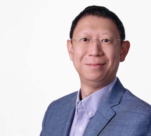 Headshot of a man with short dark hair and glasses, smiling against a white background. He is wearing a light purple button-down shirt and a blue textured blazer. The lighting is bright and even, creating a professional and friendly appearance.